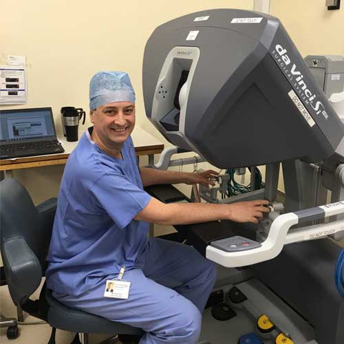 Mr Liviu Titu is wearing blue medical equipment and sits on a special chair in front of the Da Vinci Robo Surgeon command. The Robot Surgeon is big, has a display, joysticks and pedals. Mr Liviu Titu has a friendly smile on his face and looks confident in operating the machinery.
